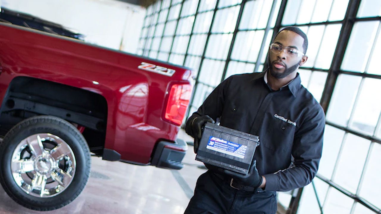 A service technician carrying a car battery past a red pickup truck