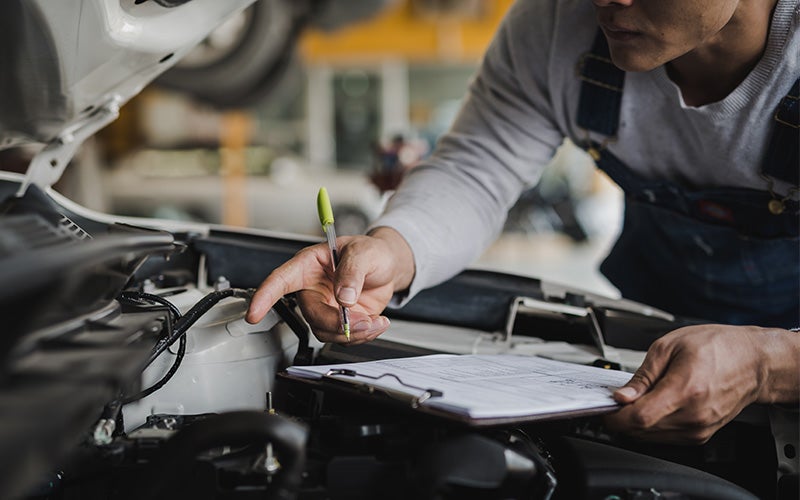 A mechanic holding a clipboard inspects an open car engine in a garage